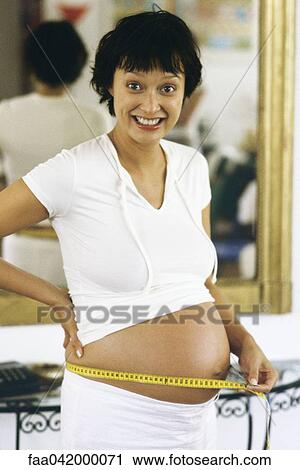 Stock Image - "Pregnant woman measuring stomach with measuring tape, smiling at camera". Fotosearch