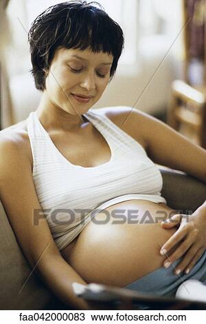 Stock Image - "Pregnant woman sitting on sofa, touching her stomach". Fotosearch