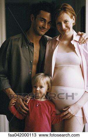 Stock Image - "Pregnant woman standing with husband and daughter, smiling at camera". Fotosearch