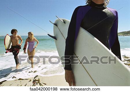 Stock Photo - "Surfer carrying board under arm, couple emerging from water together". Fotosearch