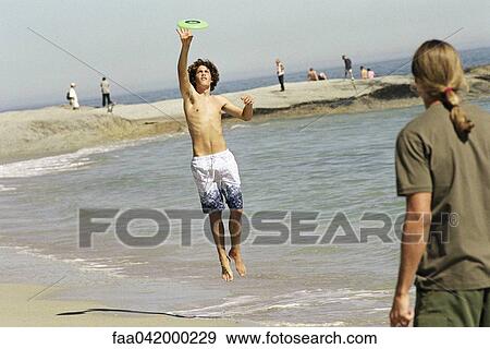 Stock Photo - "Two friends on beach throwing frisbee, one jumping to catch". Fotosearch