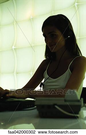 Stock Image - "Woman at desk wearing headset, using computer, smiling". Fotosearch