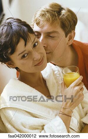 Stock Image - "Woman holding glass of orange juice, man kissing her cheek". Fotosearch