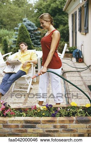 Stock Photograph - "Woman watering flowers on patio, husband relaxing with newspaper in background". Fotosearch
