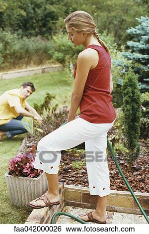 "Woman watering garden with hose, husband crouching in background" View Large Photo Image Stock Photograph - "Woman watering garden with hose, husband crouching in background". Fotosearch