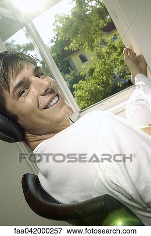 Stock Photo - "Young man listening to headphones, sitting by window with feet up". Fotosearch