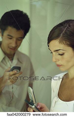 Stock Photo - "Young woman looking at cell phone, man in background lookng at own phone, smiling". Fotosearch