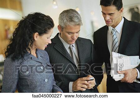Stock Photo - Business associates standing together, looking at cell phone. Fotosearch