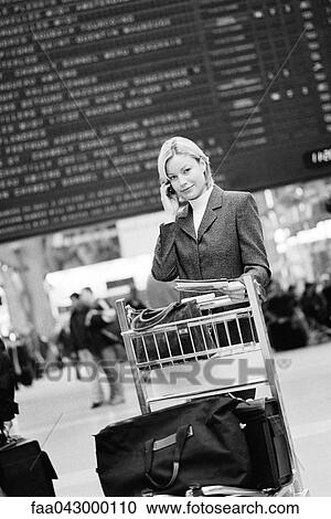 Stock Image - Business traveler phoning standing by luggage cart, arrival and departure board in background. Fotosearch