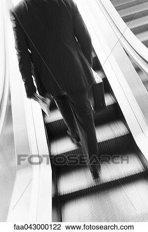 Stock Image - Businessman ascending escalator, rear view. Fotosearch