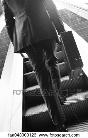 Stock Image - Businessman carrying briefcase ascending escalator, rear view. Fotosearch
