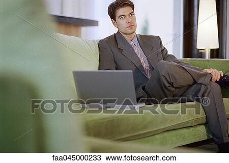 Stock Image - Businessman sitting on sofa with laptop, looking away. Fotosearch