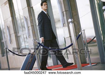 Stock Photo - Businessman walking toward building entrance, pulling suitcase behind him. Fotosearch