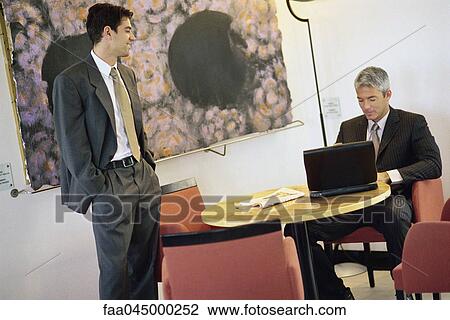 Stock Image - Businessmen chatting in break room, one using laptop computer. Fotosearch
