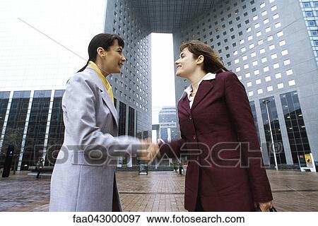 Stock Photo - Businesswomen shaking hands, meeting in plaza outside office building. Fotosearch