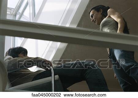 Stock Image - Couple in conversation, man seated in armchair, woman standing, low angle view. Fotosearch