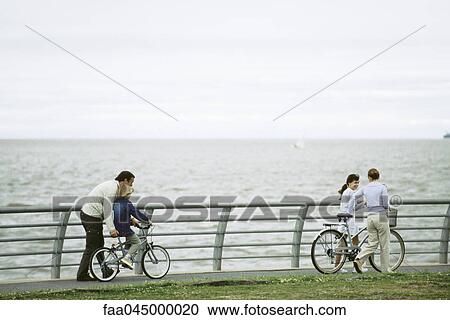 Stock Image - Family riding bicycles together at seaside park, father helping son learn to ride. Fotosearch