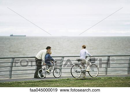 Stock Photo - Father helping son learn to ride bicycle, mother watching. Fotosearch