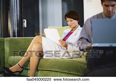 Stock Photography - Female executive reading documents on sofa, man working on laptop in foreground. Fotosearch