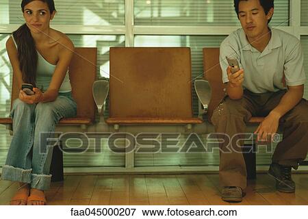 Stock Photo - Man and woman sitting in waiting room, holding cell phones, woman glancing sideways at man. Fotosearch