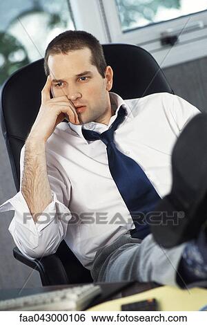 Stock Photograph - Man with feet up on office desk holding head looking away. Fotosearch