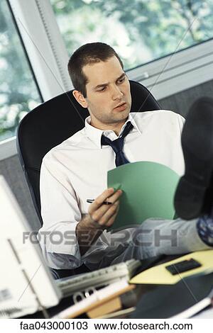Stock Image - Man with feet up on office desk reviewing document. Fotosearch