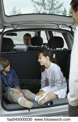 Stock Image - Siblings sitting in back of station wagon, parents watching. Fotosearch