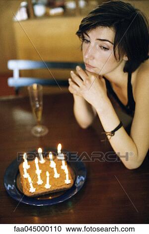 Stock Image - Woman at table with birthday cake looking away wistfully. Fotosearch