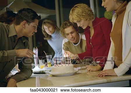 Stock Image - Woman lighting candles on birthday cake, others standing by watching. Fotosearch