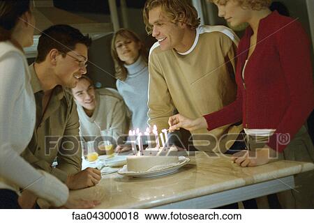 Stock Photo - Woman lighting candles on birthday cake, friends standing nearby smiling, watching. Fotosearch