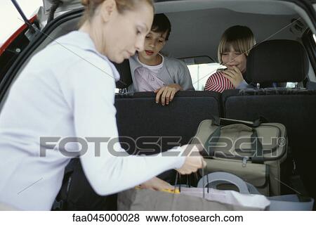 Stock Photo - Woman organizing luggage in back of car, children watching from back seat. Fotosearch