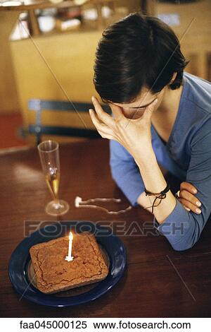 Stock Photography - Woman sitting at table alone with anniversary cake, covering eyes. Fotosearch