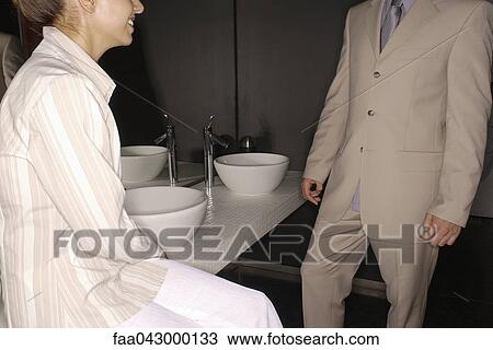 Stock Image - Woman sitting on bathroom sink counter edge smiling at well dressed man. Fotosearch