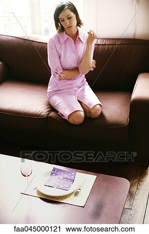 Stock Image - Woman sitting on sofa looking down a place setting set on coffee table, high angle view. Fotosearch