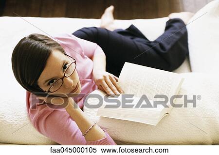 Stock Photography - Woman sitting on sofa reading book, looking up at camera, high angle view. Fotosearch