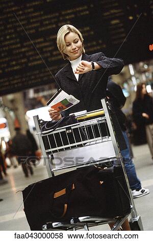Stock Photo - Woman standing with luggage cart in train station holding travel ticket looking at watch. Fotosearch