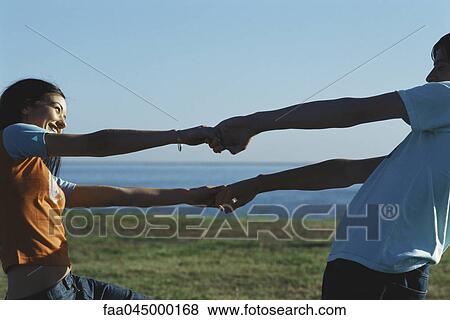 Stock Photo - Young couple facing one another holding hands and leaning back smiling. Fotosearch