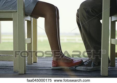 Stock Photography - Young couple seated at table facing one another, woman's feet touching mans, low section. Fotosearch