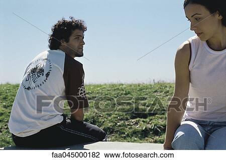 Stock Image - Young couple sitting apart on park bench, glancing over shoulders at one another. Fotosearch