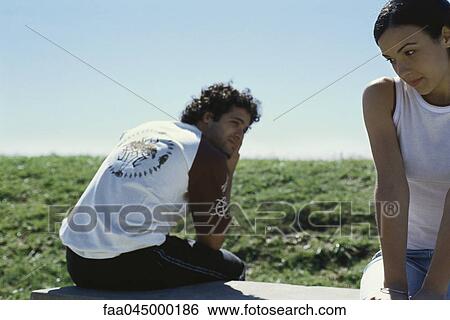 Stock Photograph - Young man and woman sitting on bench, man glancing sideways over shoulder at woman. Fotosearch