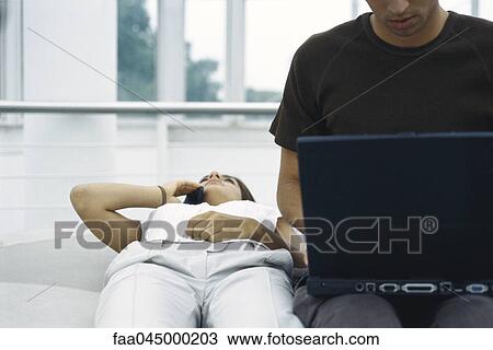 Stock Image - Young woman lying on back using cell phone, man using laptop beside her. Fotosearch