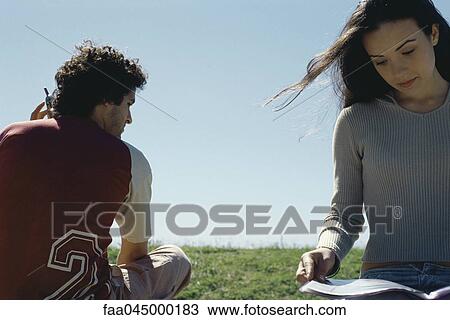 Stock Image - Young woman sitting outdoors reading magazine, man using cell phone behind her. Fotosearch