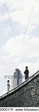 Stock Image - Business professionals standing on wall with arms folded, skyscraper in background. Fotosearch