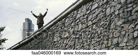 Businessman standing on wall with arms raised, skyscraper in background View Large Photo Image Stock Photo - Businessman standing on wall with arms raised, skyscraper in background. Fotosearch