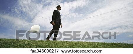 Stock Photo - Businessman walking barefoot in grassy field, empty chair nearby. Fotosearch