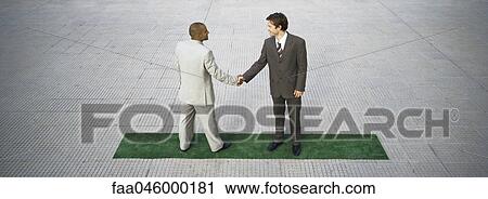 Stock Image - Businessmen standing on strip of artificial turf shaking hands. Fotosearch