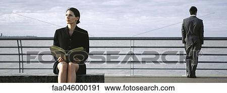 Stock Image - Businesswoman on bench at waterfront with open book on lap, man at railing looking at view in background. Fotosearch