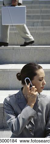 Businesswoman using cell phone outdoors, person walking on steps in background View Large Photo Image Picture - Businesswoman using cell phone outdoors, person walking on steps in background. Fotosearch