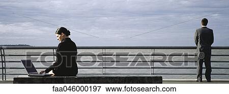 Businesswomen on waterfront bench using laptop computer, man at nearby railing looking at view View Large Photo Image Stock Photo - Businesswomen on waterfront bench using laptop computer, man at nearby railing looking at view. Fotosearch