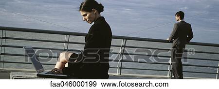 Stock Photo - Businesswomen on waterfront bench using laptop computer, man at nearby railing looking at view. Fotosearch